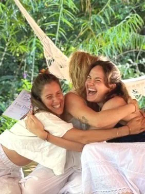 Two women hugging and smiling on a hammock in a tropical setting.