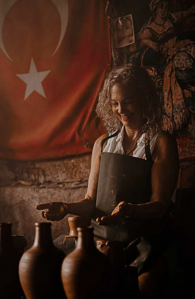 A woman with curly gray hair and a white sleeveless shirt, wearing a black apron, is working with clay pottery in a dimly lit room. There are several vases in the foreground. A Turkish flag is hanging on the wall behind her, along with fabric art and