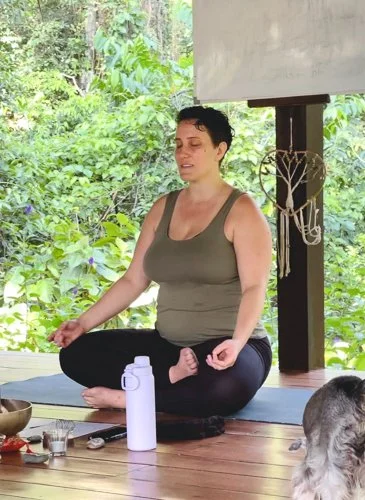 A woman practicing meditation outdoors on a wooden deck surrounded by greenery, sitting cross-legged on a mat with her hands resting on her knees.