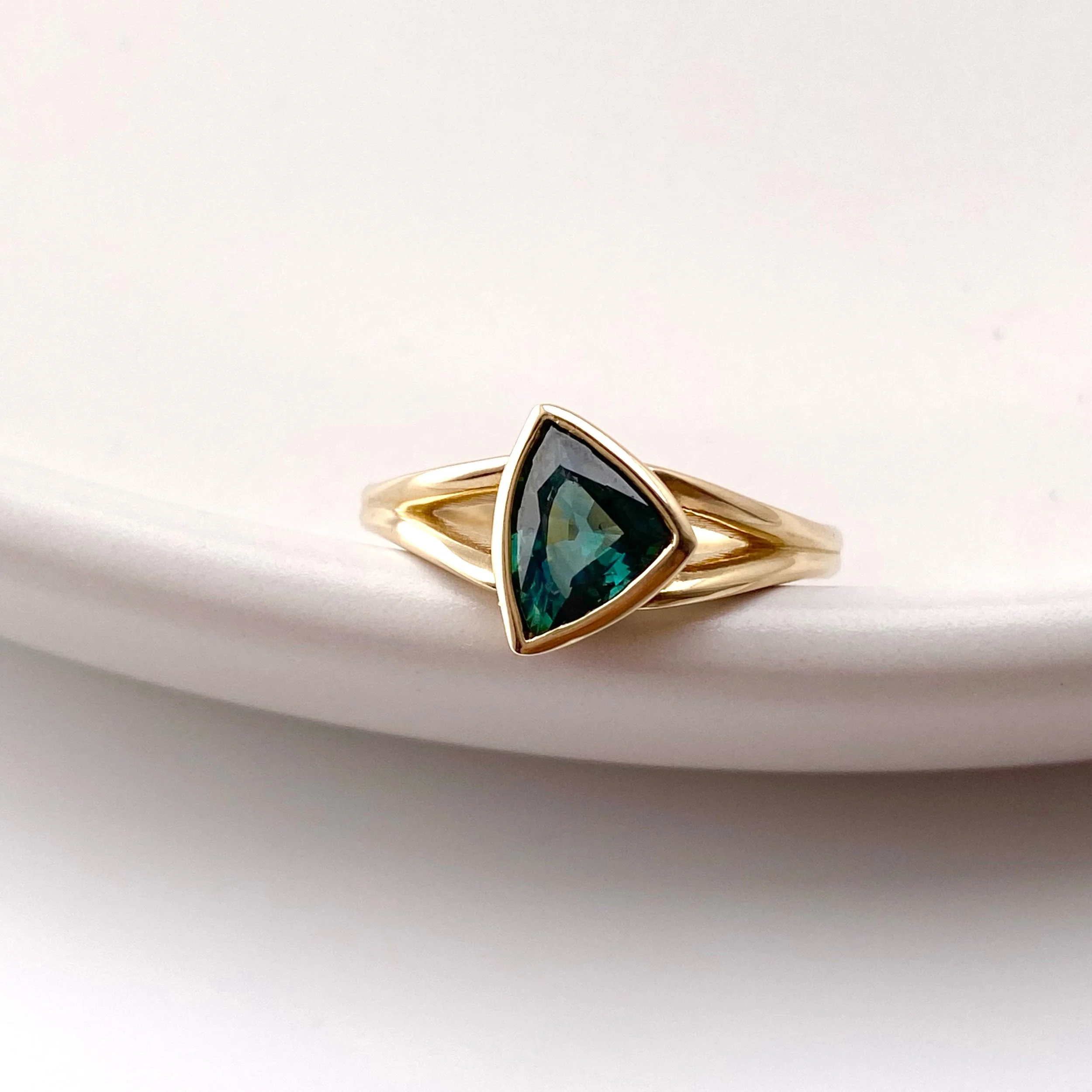 modern trillion dark teal sapphire bezel set split shank engagement ring 14k yellow gold on dish Kim Drosdick Jewellery Toronto