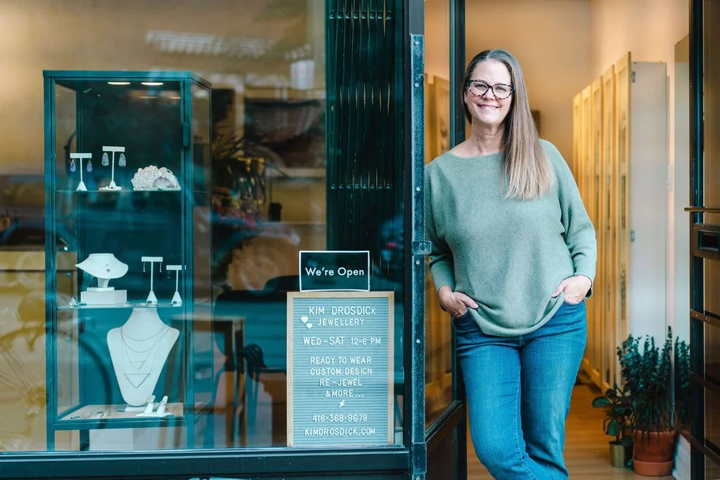 A woman with long hair and glasses standing outside a jewelry store, smiling.