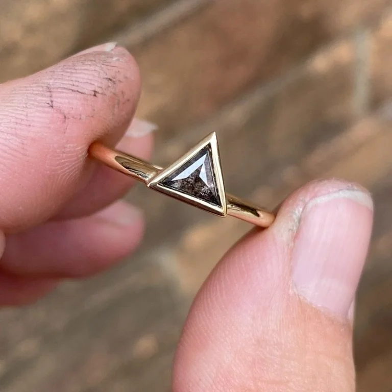 darker triangle salt and pepper diamond bezel set off side bottom justified to round wire band 14k yellow gold held between 2 fingers front view Kim Drosdick Jewellery Toronto