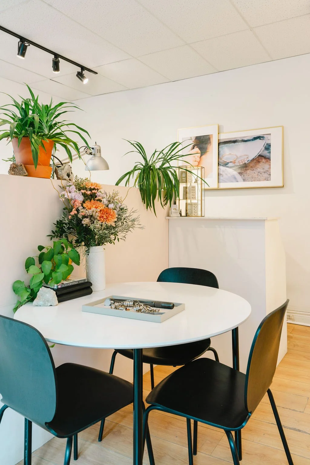 A cozy interior space with a white round table surrounded by black chairs, decorated with a tray of jewelry. Behind the table is a wall with framed artwork, potted plants, and a shelf displaying small objects and plants.