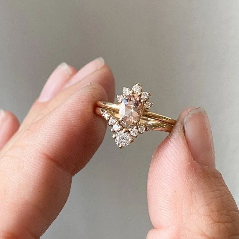 peachy pink oval morganite with little crown of Canadian diamonds claw set in 14k yellow gold narrow rounded band held in hand with accented wedding ring Kim Drosdick Jewellery Toronto