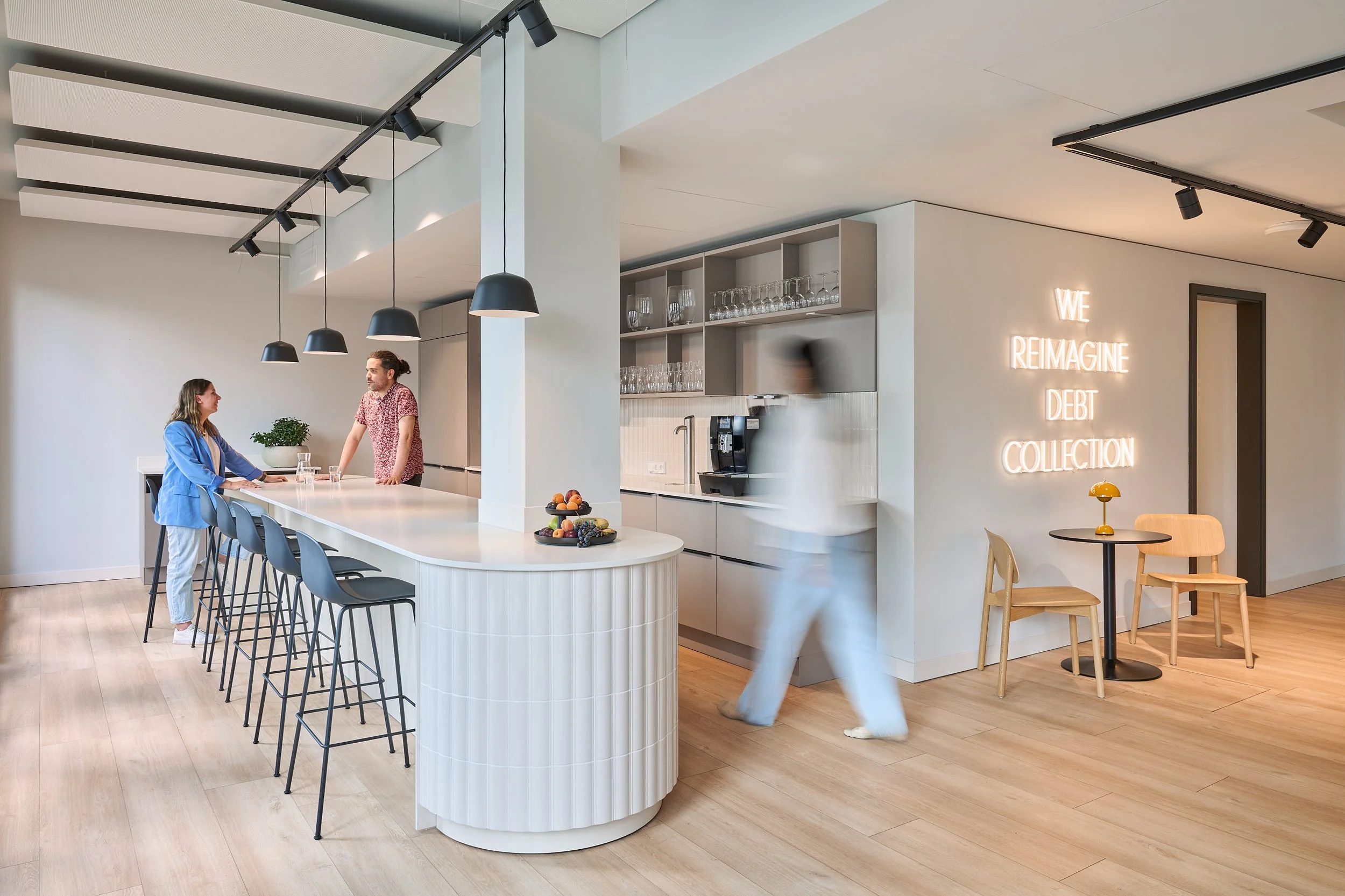 Büroküche als sozialer Treffpunkt, weiß geflieste Kücheninsel, hellgrauer Wandanstrich, hellgraue Küchenkorpen, heller Holzfußboden, Neon Schriftzug