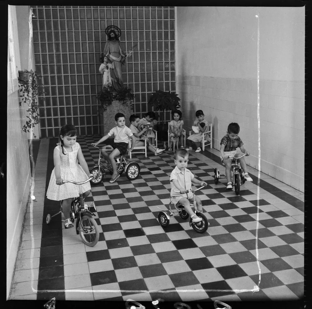 Centro Maternal de la Cárcel de Mujeres [Interior de una cárcel de mujeres, sala presidida por una escultura de San José con el Niño, pavimento ajedrezado y paredes con zócalo azulejado, un grupo de niños montan en triciclo en el interior mientras otros. fotografía: pando barrero
