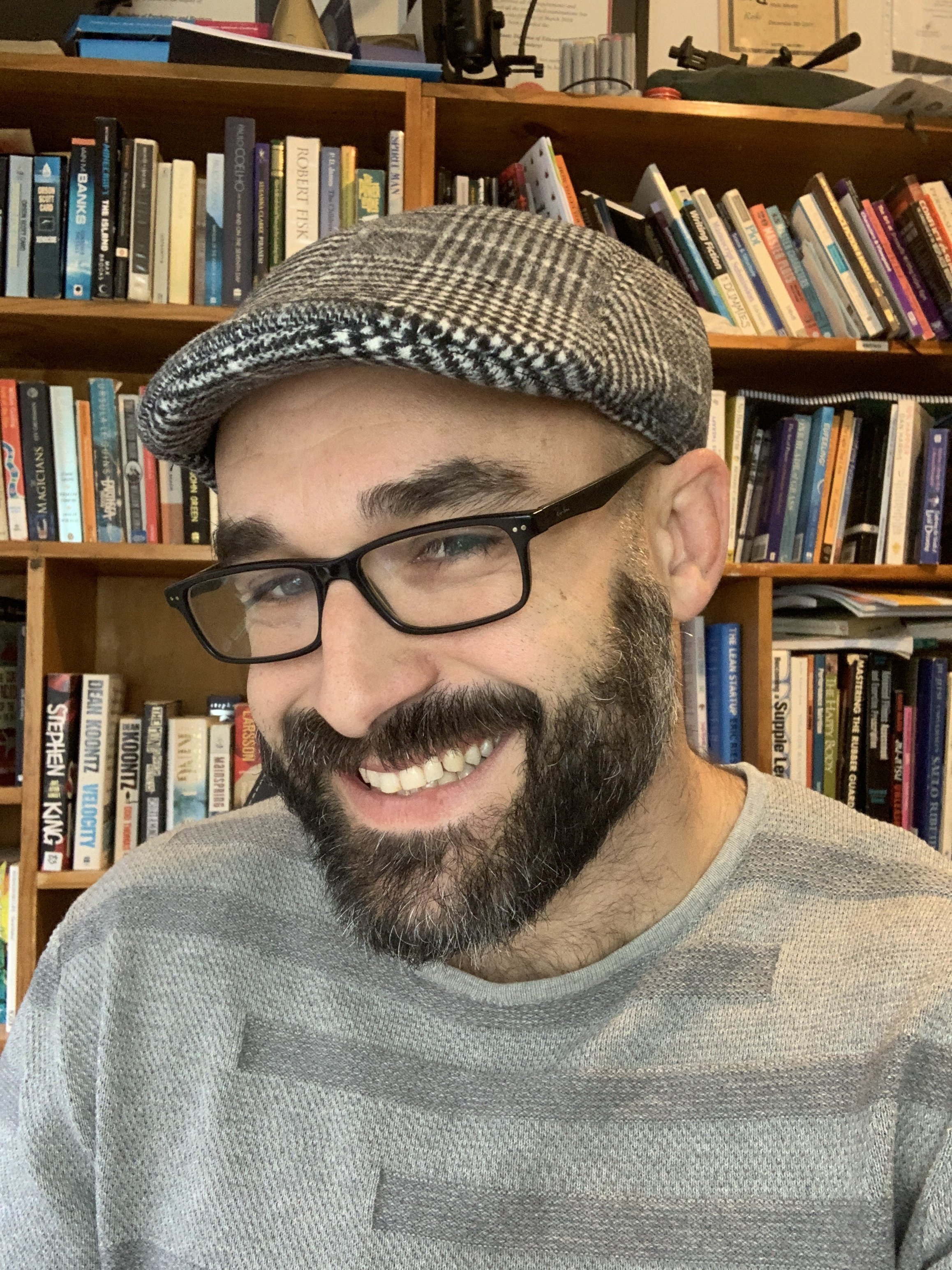 A smiling man with glasses and a beard wearing a checkered flat cap and a gray textured sweater, standing in front of a bookshelf filled with books.