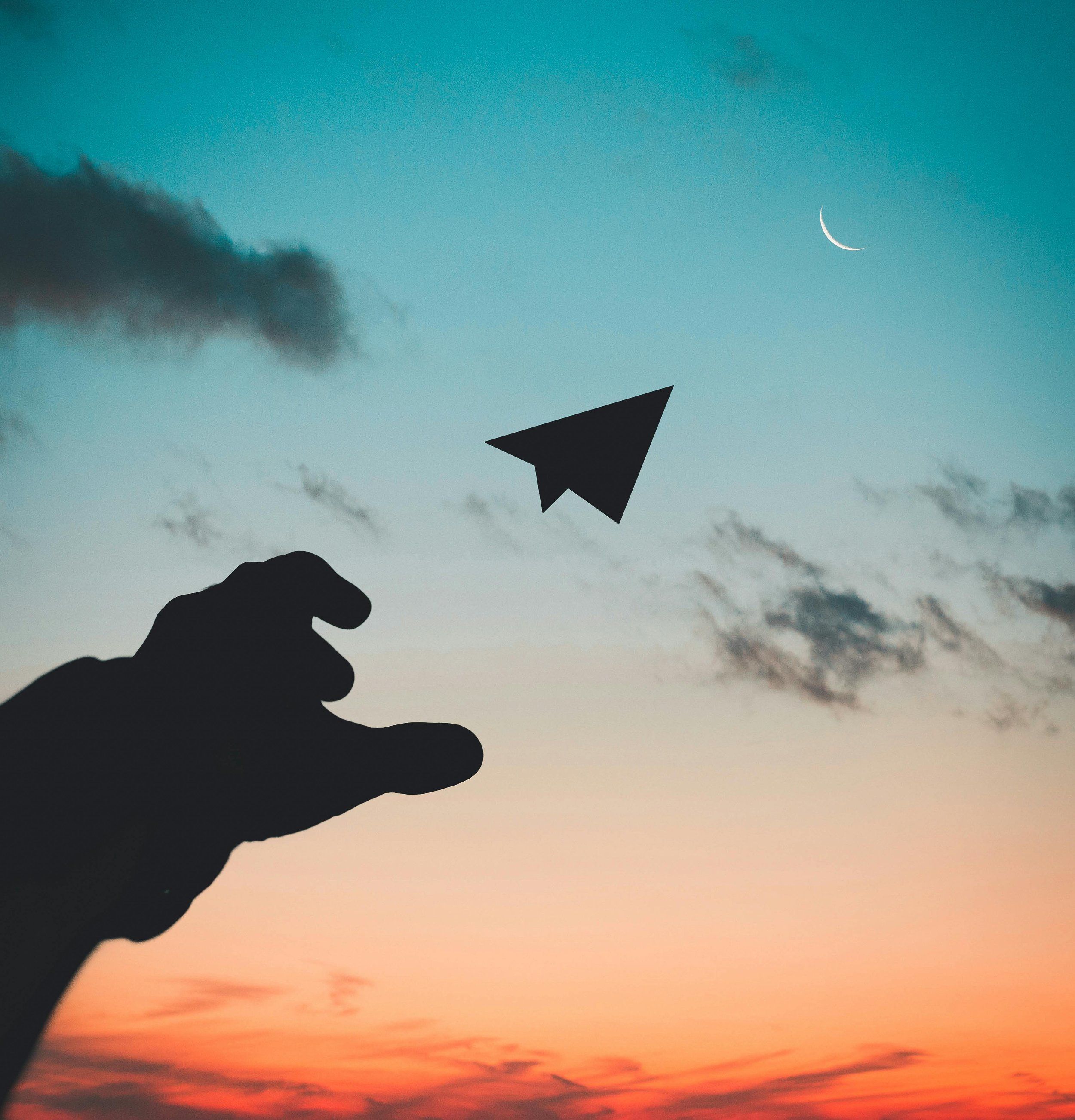 Silhouette of a hand and paper airplane against a sunset sky with crescent moon.