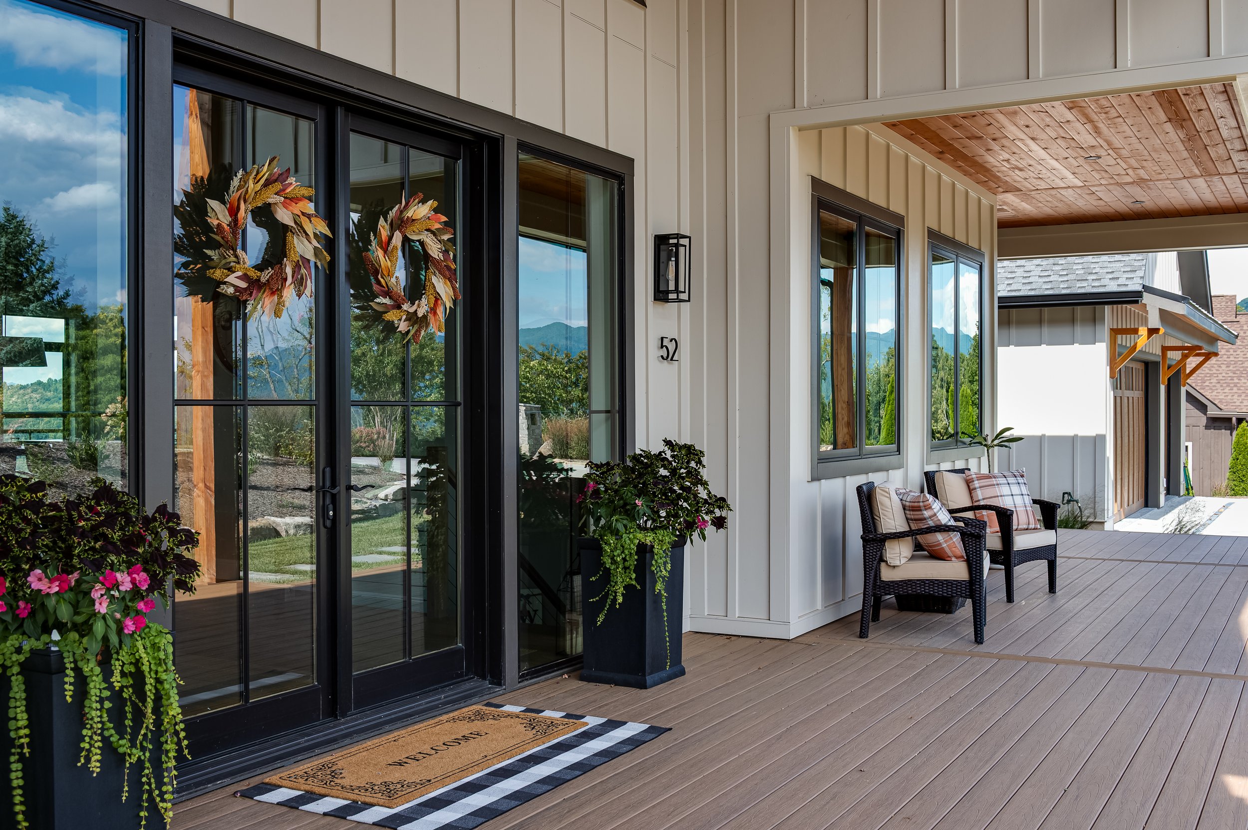 A covered outdoor porch area with sliding glass doors decorated with fall-themed wreaths. There are flower pots with pink and green plants, wicker chairs with cushions, and brown wooden flooring. The porch has beige siding, a wooden ceiling, and a mo