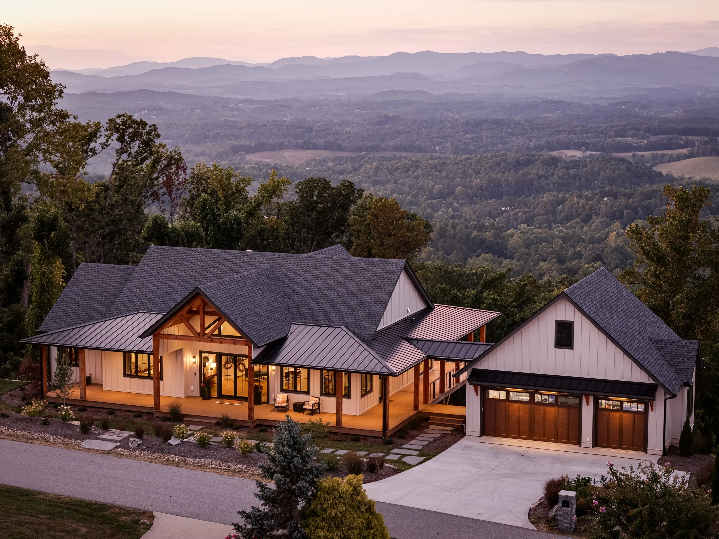 A modern house with large windows and a metal roof, situated in a hilly landscape with trees and mountains in the background, taken during dusk.