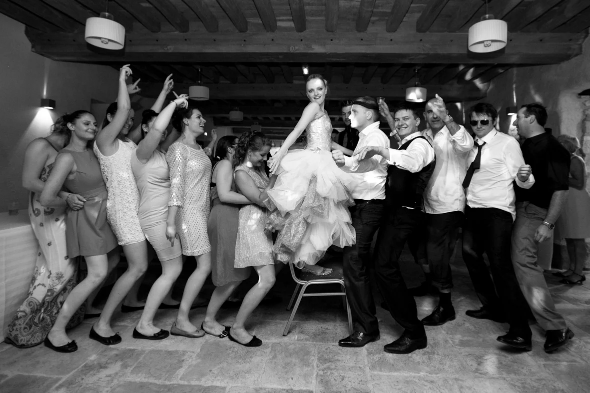 Group of people at a celebration, with a woman in a dress being lifted on a chair by men dressed in shirts and ties, surrounded by women in dresses, all smiling and enjoying the event in a decorated indoor venue.