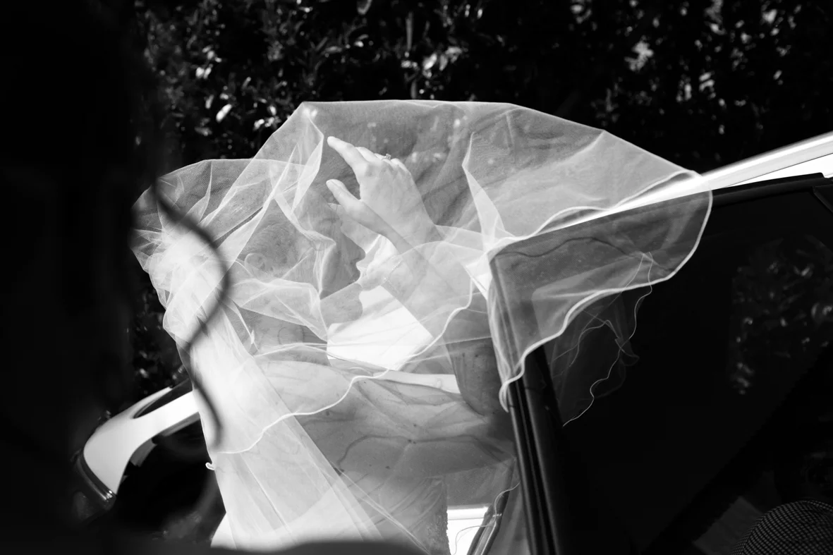 A person dressed in white with a veil over their head, sitting in a car, reaching out with their hand.