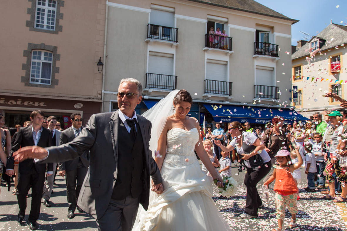 A wedding celebration on a city street with a bride and groom walking hand-in-hand, surrounded by guests throwing confetti and children dancing, with onlookers and buildings in the background.