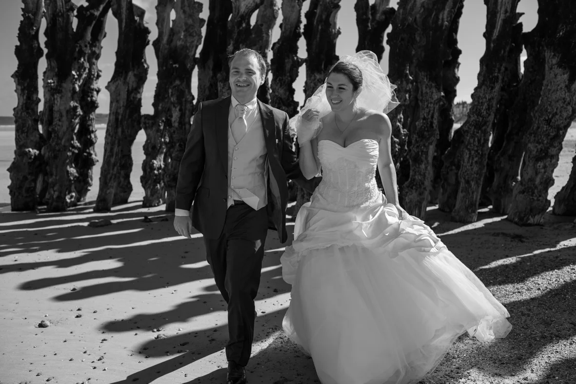 Un couple de mariés marche sur la plage, souriant, avec des structures en bois derrière eux.