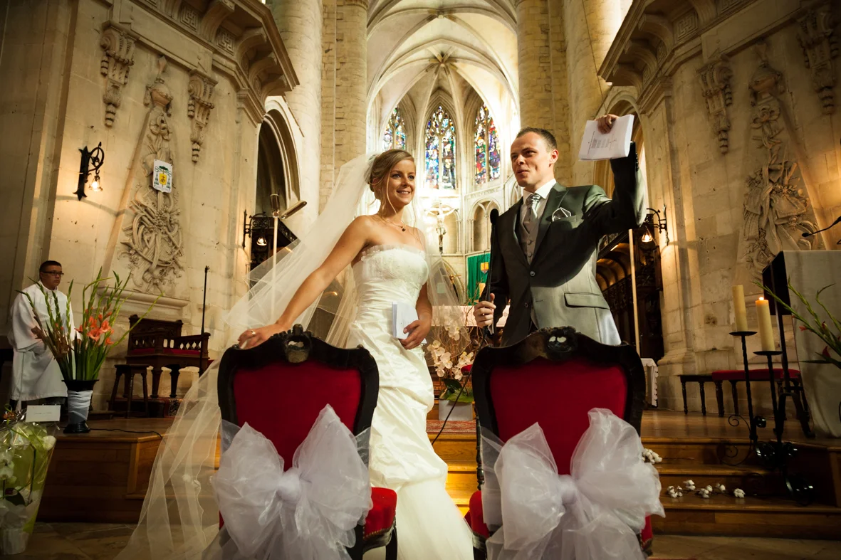 Bride and groom stand at the altar during their wedding ceremony inside a church with stained glass windows in the background. The bride is wearing a white strapless wedding gown and veil, smiling. The groom is wearing a suit and tie, holding a paper