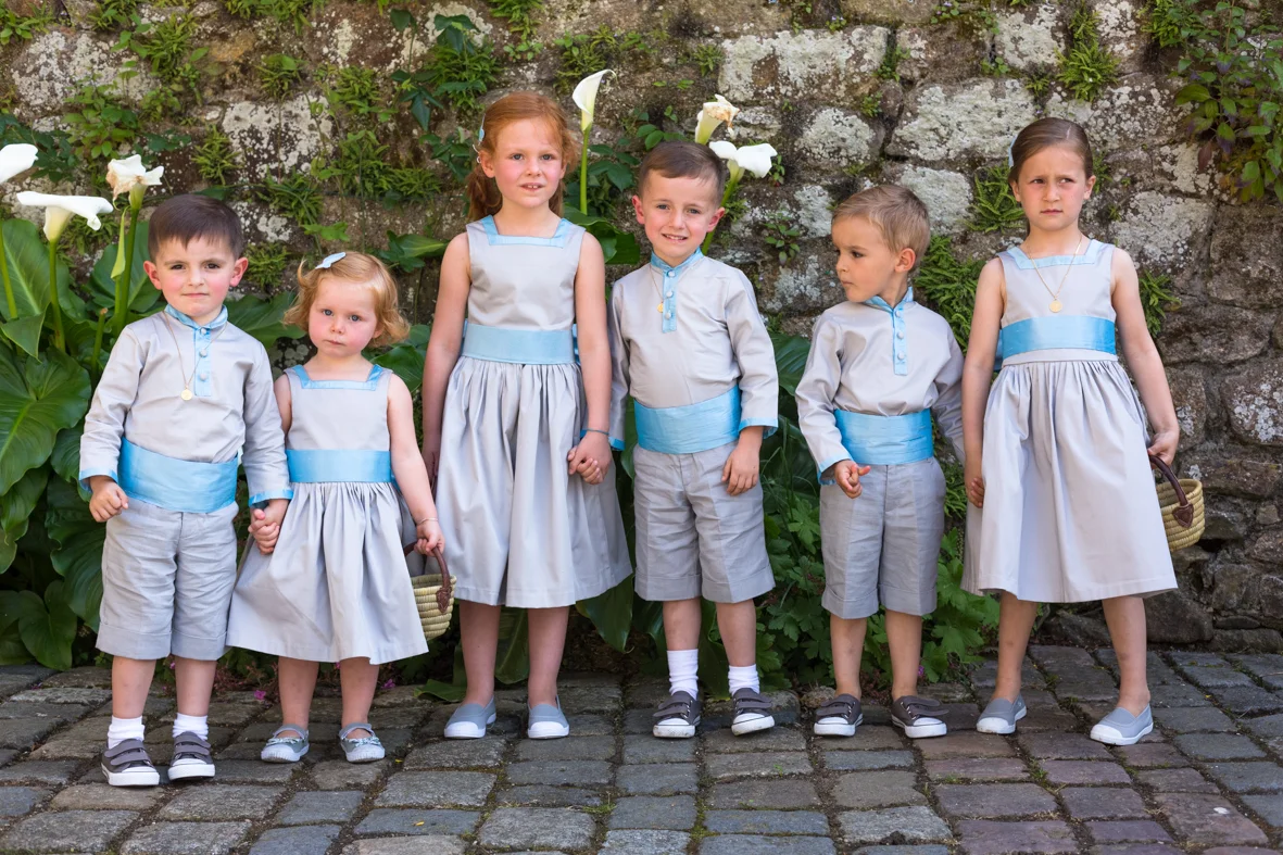 Groupe de sept enfants habillés en vêtements coordonnés, posant devant un mur en pierre avec des plantes vertes en arrière-plan.