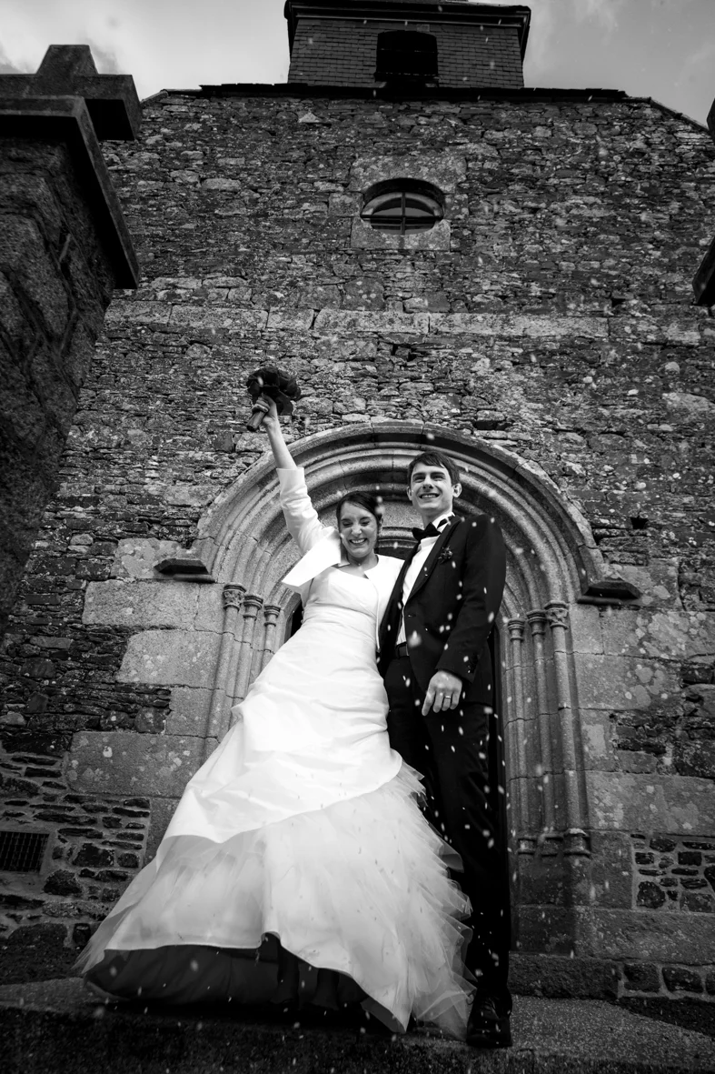 Black and white photo of a happy couple in wedding attire celebrating outside a stone church. The bride is holding a trophy high, and confetti is falling around them.