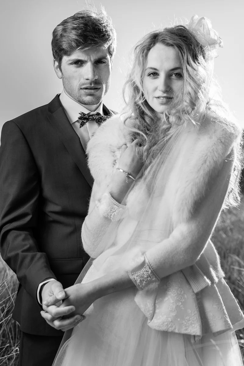 Une photo en noir et blanc d'un homme et d'une femme posant ensemble, semblant être lors d'une séance photo. L'homme porte un costume avec un nœud papillon, tandis que la femme porte une robe et une veste en fourrure avec un accessoire dans ses cheve