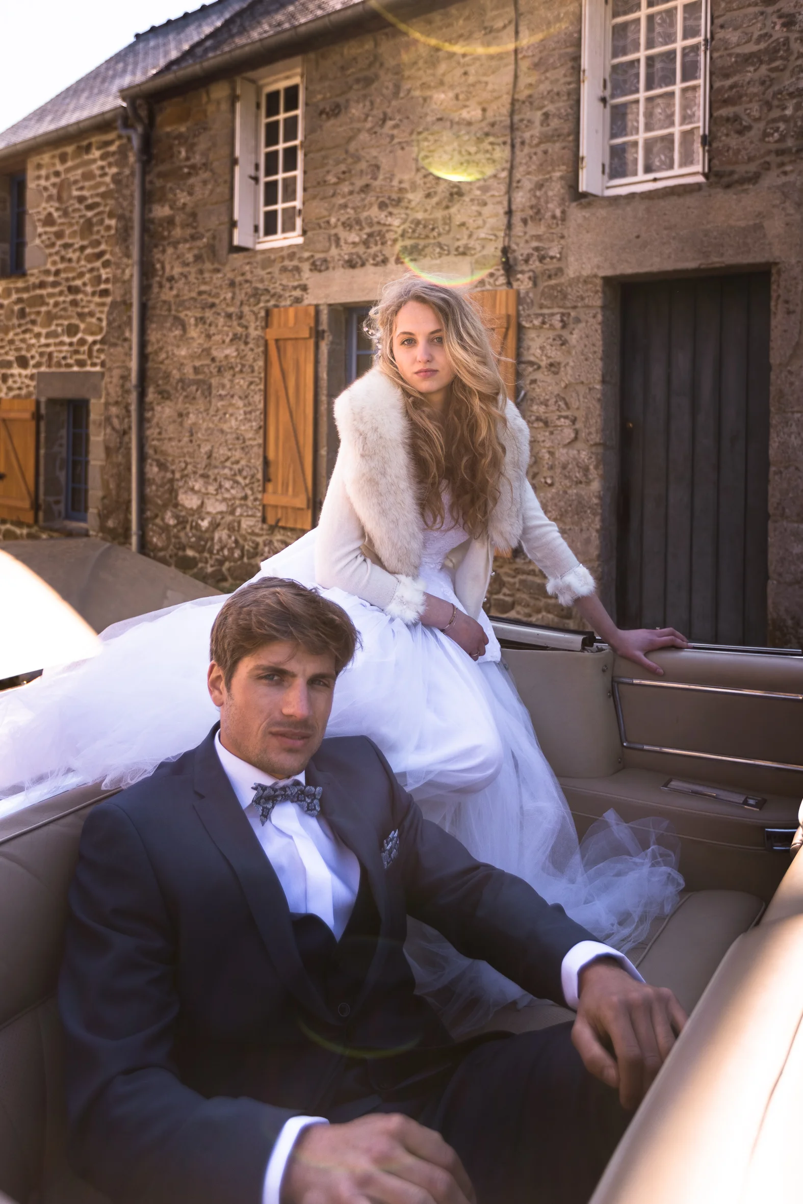 A young woman in a white dress and fur stole sitting on the back of a vintage car, with a young man in a dark suit and bow tie sitting in the driver's seat. They are outside in front of a stone building with wooden shutters.