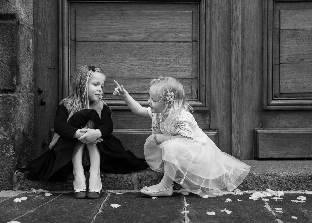 Two young girls dressed in formal dresses sitting on a stone step outside a wooden door, with one girl pointing her finger at the other.