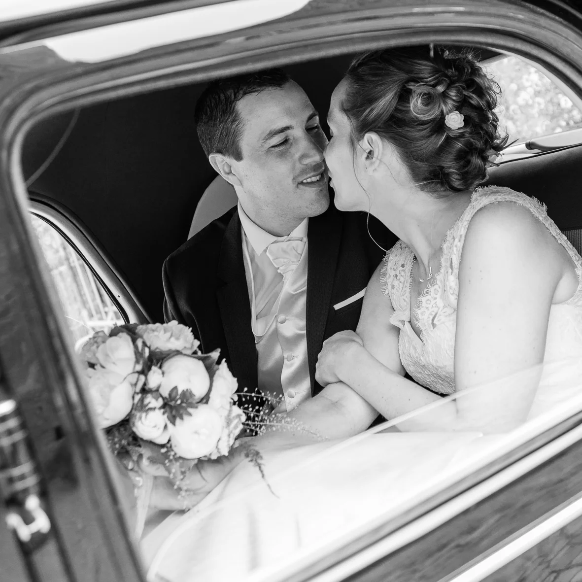 Black and white photo of a newlywed couple sitting in a vintage car, sharing a close moment. The woman is in a wedding dress with lace details, and the man is in a tuxedo. A bridal bouquet is visible in the foreground.