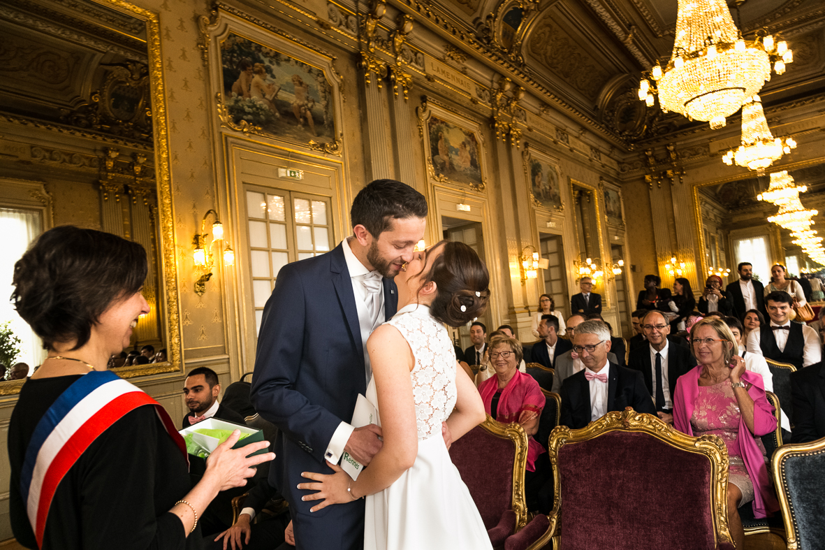 A wedding ceremony taking place in an ornate, gold-accented ballroom with chandeliers, where a bride and groom are sharing a kiss. The bride is in a white lace dress, and the groom is in a navy suit. Guests dressed in formal attire are seated and smi