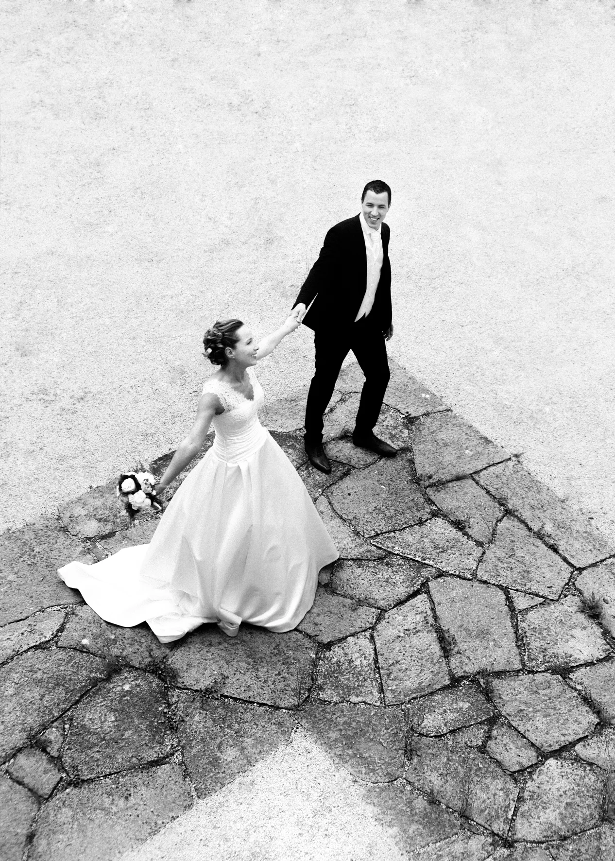 Black and white photo of a bride and groom holding hands on a cracked stone surface, looking up and smiling. The bride wears a wedding gown and holds a bouquet, while the groom wears a suit.