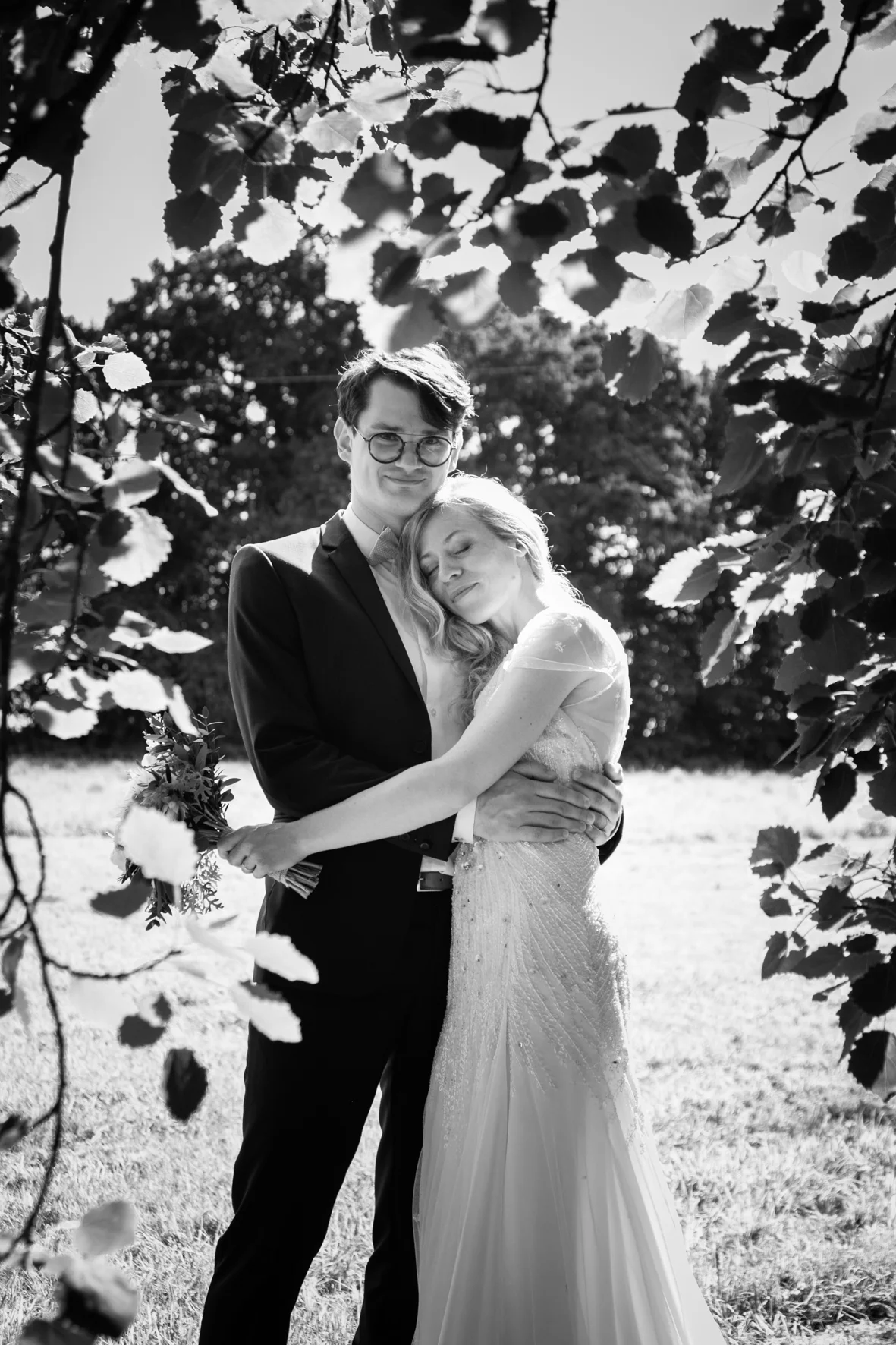 A black-and-white photo of a loving couple, dressed in wedding attire, standing outdoors. The man is wearing glasses and a suit, and the woman is in a wedding dress, embracing each other with eyes closed, surrounded by trees and foliage.