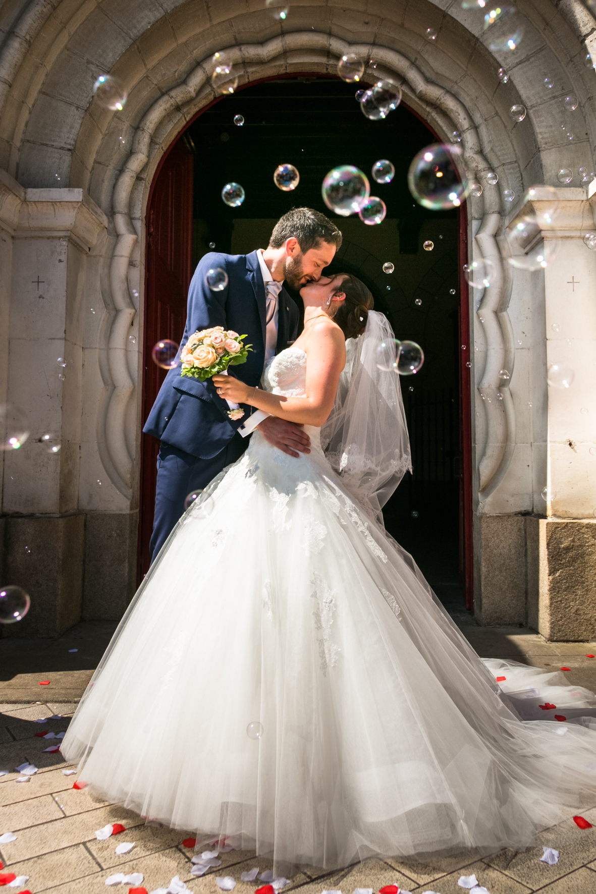 A newlywed couple sharing a kiss in front of a stone archway, surrounded by floating bubbles and scattered flower petals, with the bride wearing a white wedding gown and veil, and the groom in a blue suit holding a bouquet.