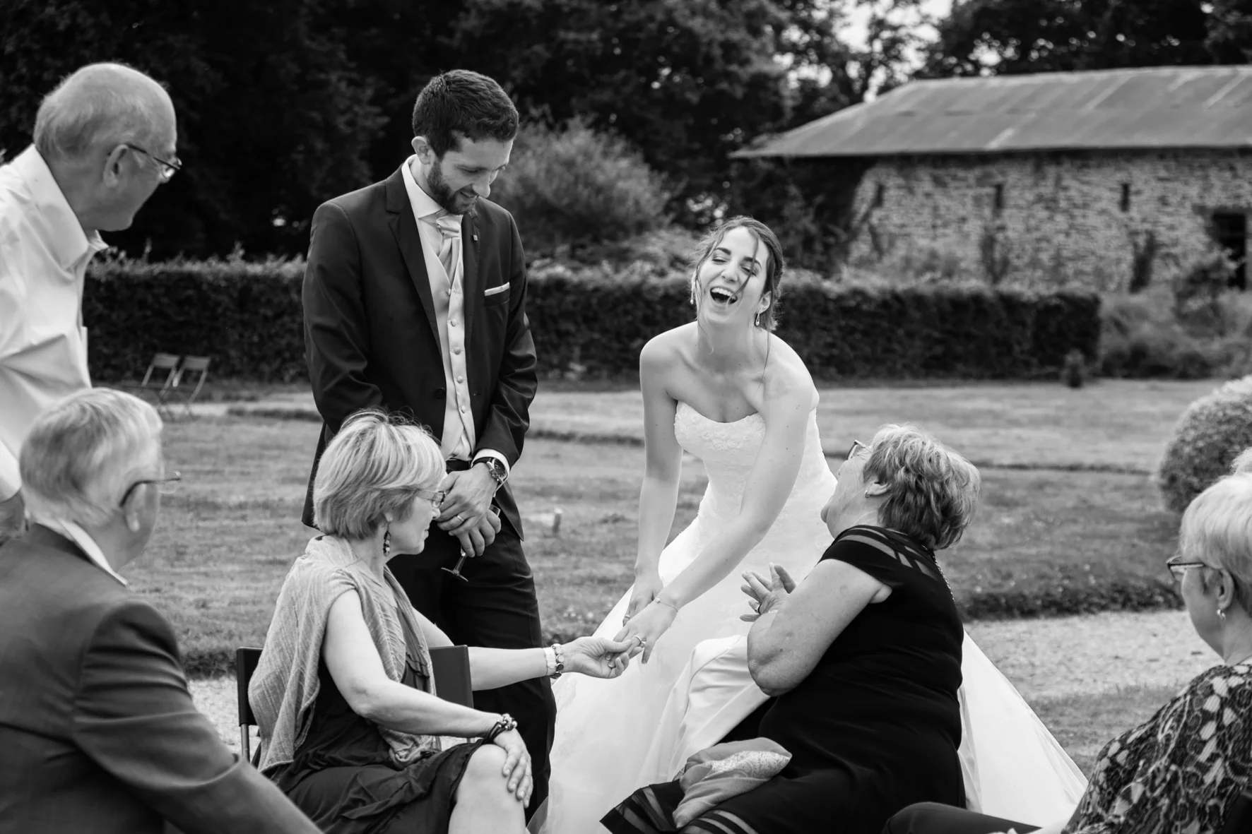 A black-and-white photo of a wedding celebration outdoors. The bride, in a strapless wedding gown, is laughing and holding hands with an older woman seated in front of her. The groom, in a dark suit, is standing nearby, smiling. Several other guests 