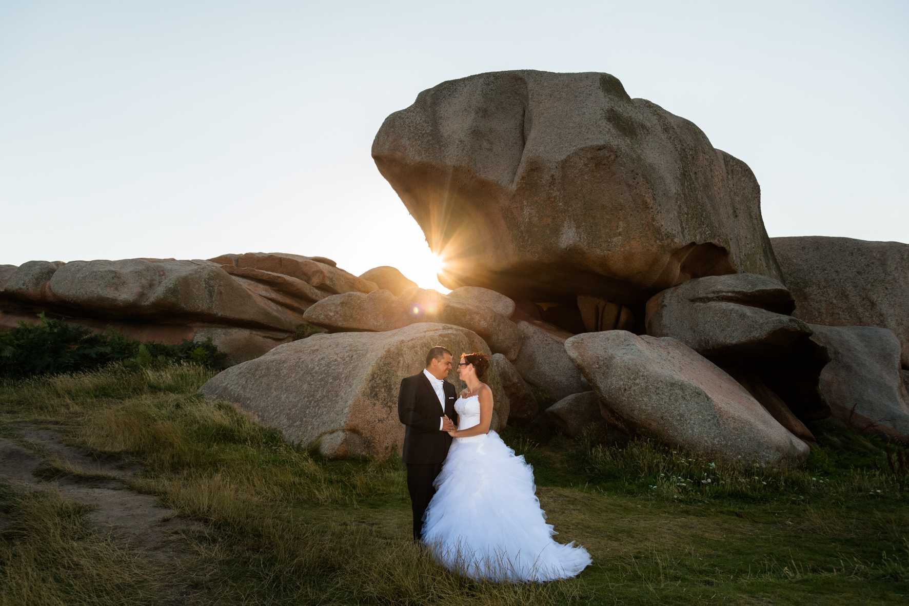 A bride and groom in wedding attire standing close together in front of large rocks during sunset in a natural landscape.