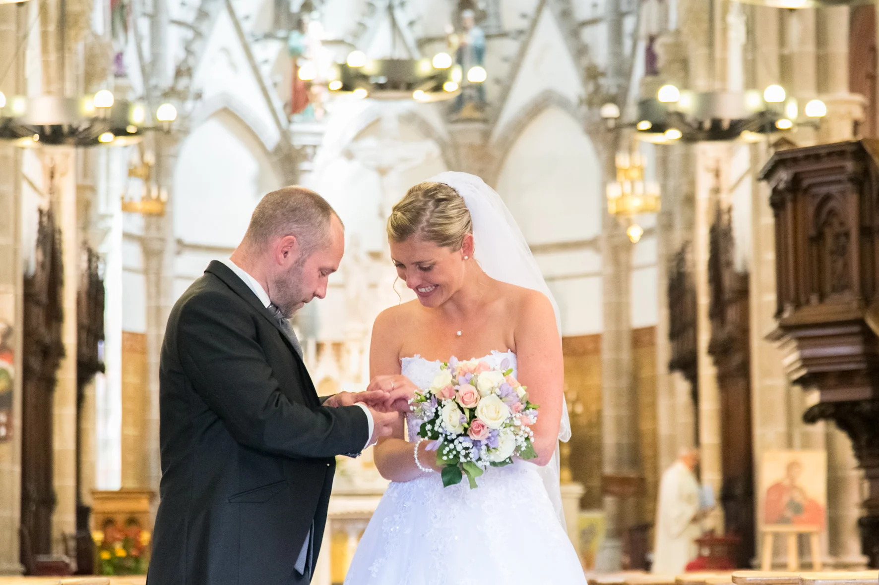 A bride and groom exchanging rings during their wedding ceremony inside a church, with the bride holding a bouquet of flowers and smiling.