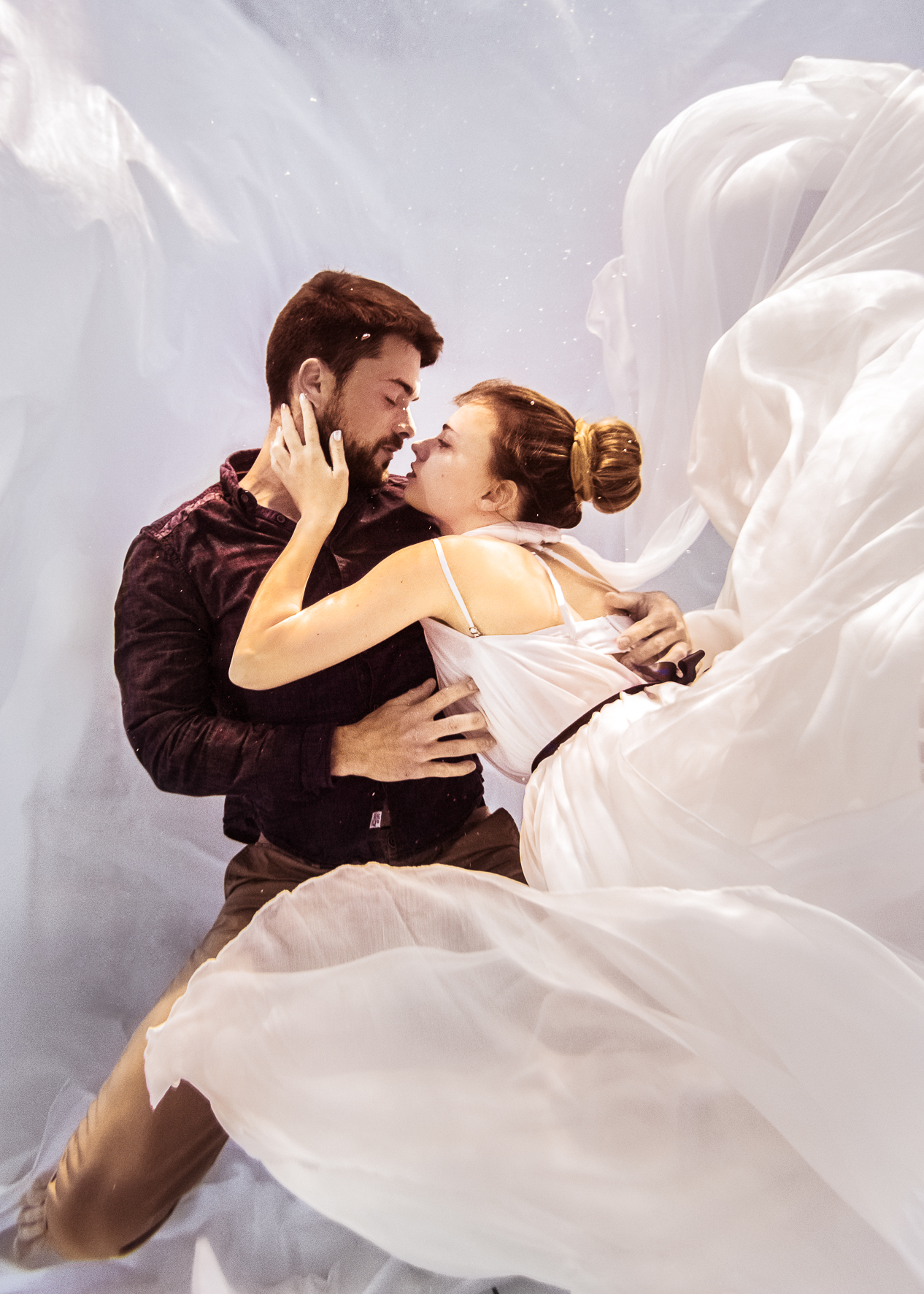 A man and woman in an intimate pose lying on white fabric with a neutral background.