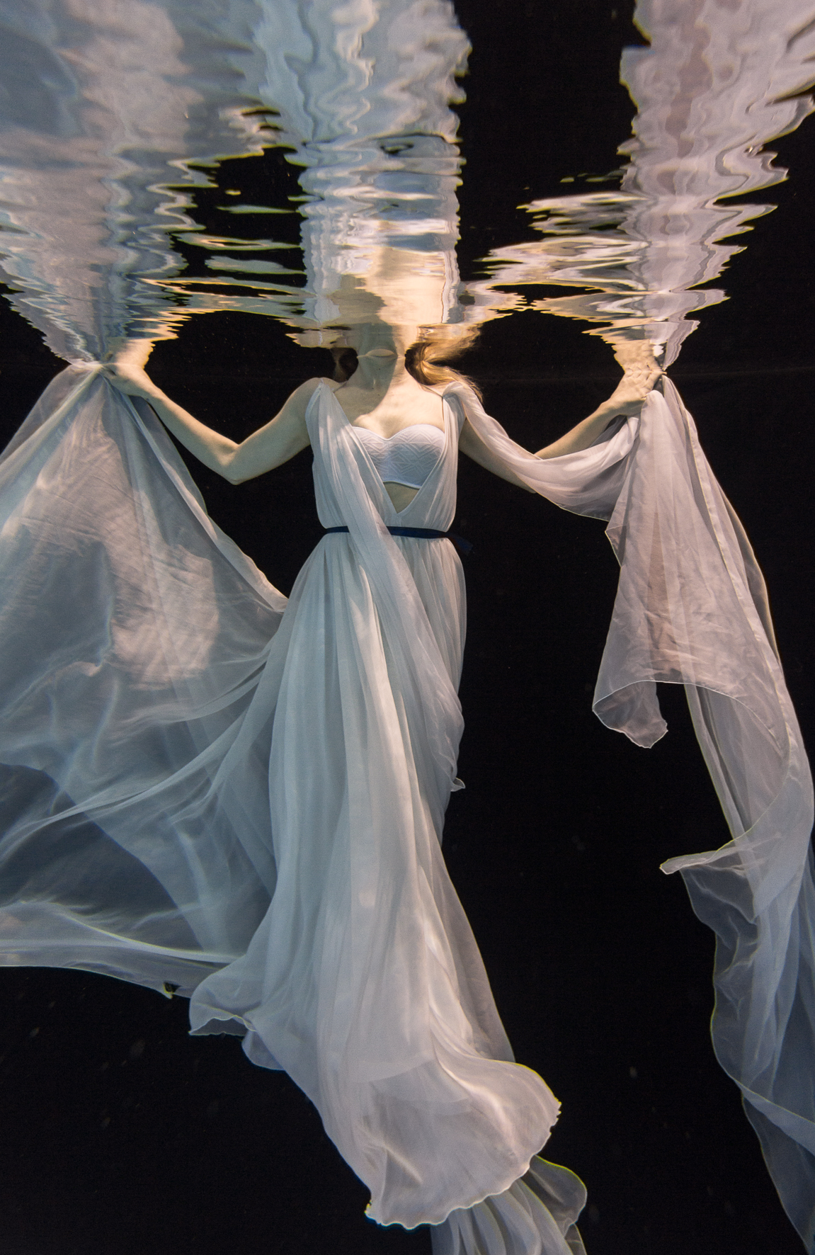 A woman in a flowing white dress is underwater, holding her dress out on both sides, with the water surface visible above her head.