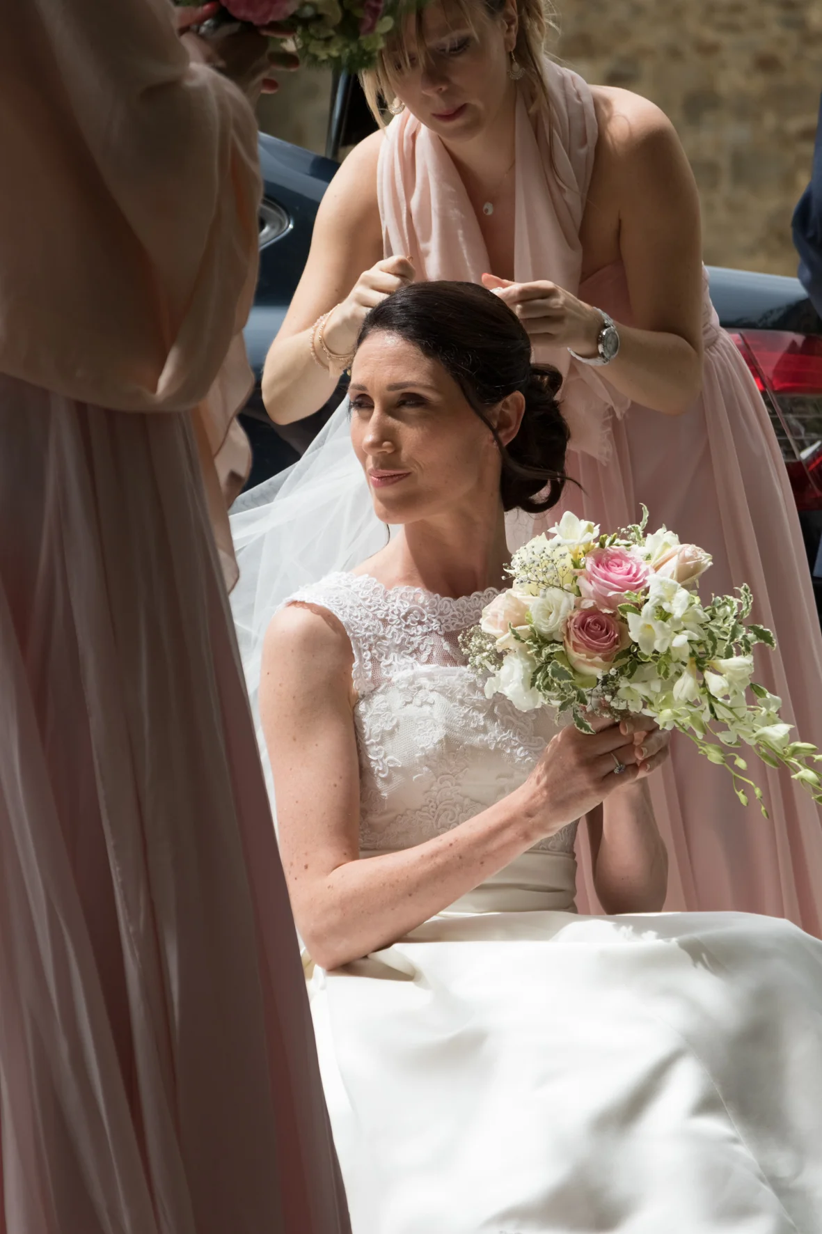 Une mariée assise, entourée de ses demoiselles d'honneur en robes roses pâles, tenant un bouquet de fleurs roses et blanches, lors d'une cérémonie de mariage en plein air.