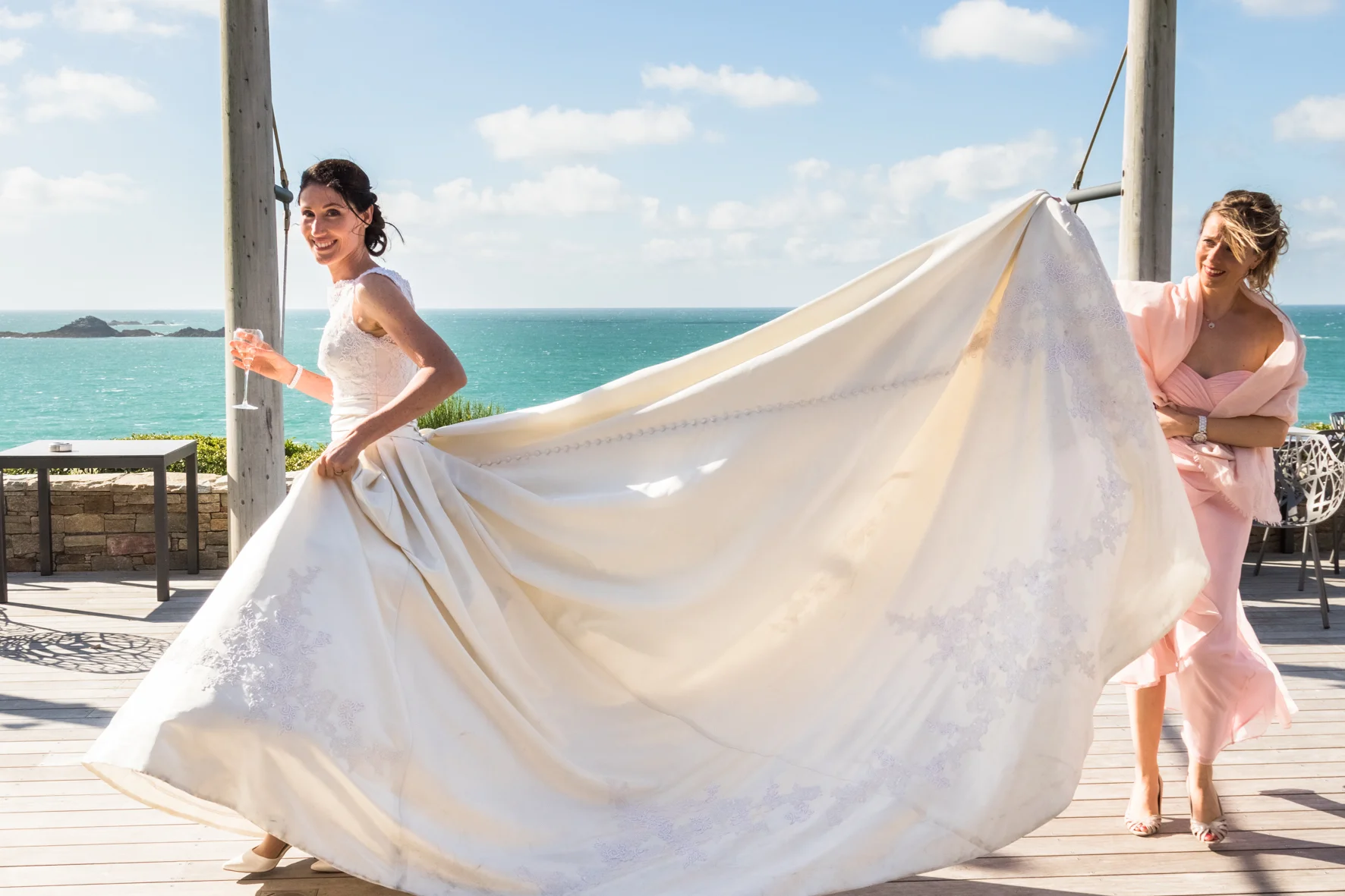 Bride in a white wedding dress holding her skirt with a woman in pink assisting, against a seaside backdrop with a deck and ocean view.