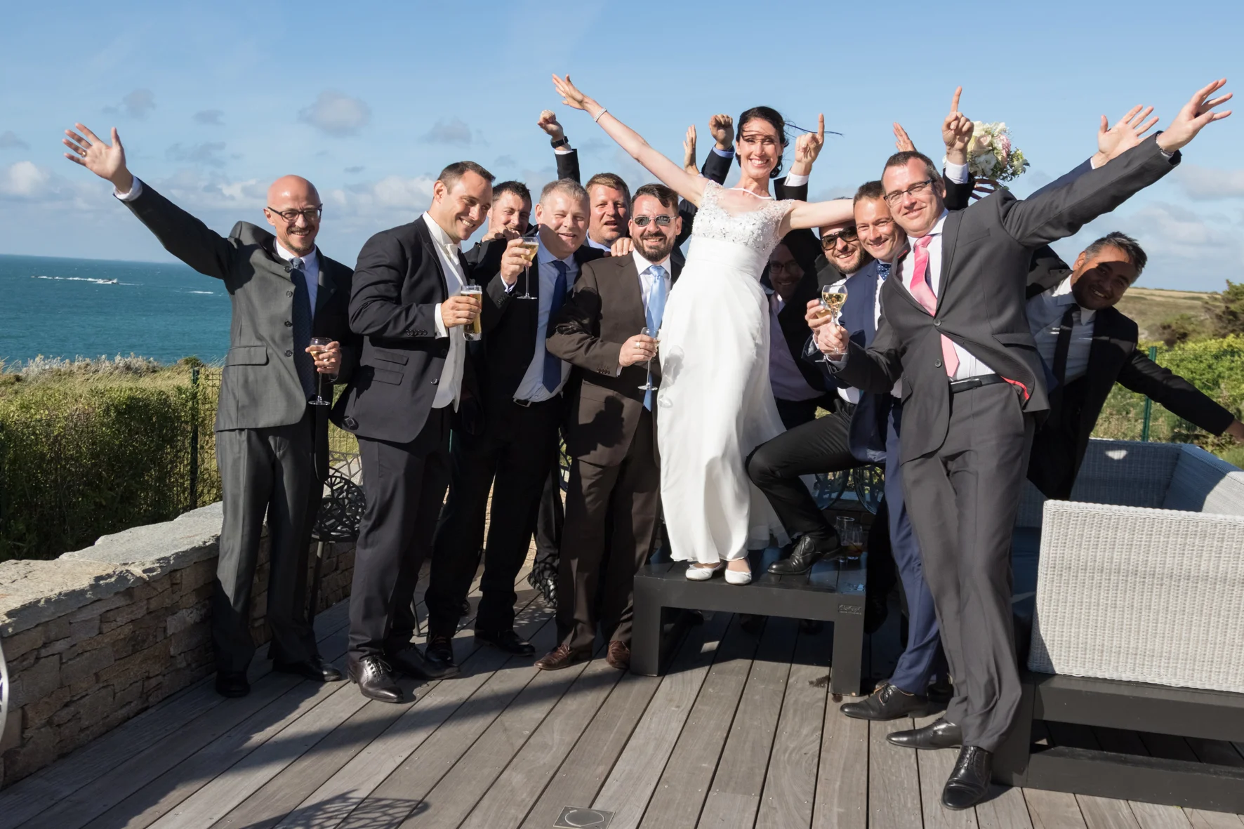 Groupe de personnes en costume, célébrant un mariage sur une terrasse avec vue sur la mer, avec la mariée en robe blanche au centre, tous souriants et tenant des verres de champagne.