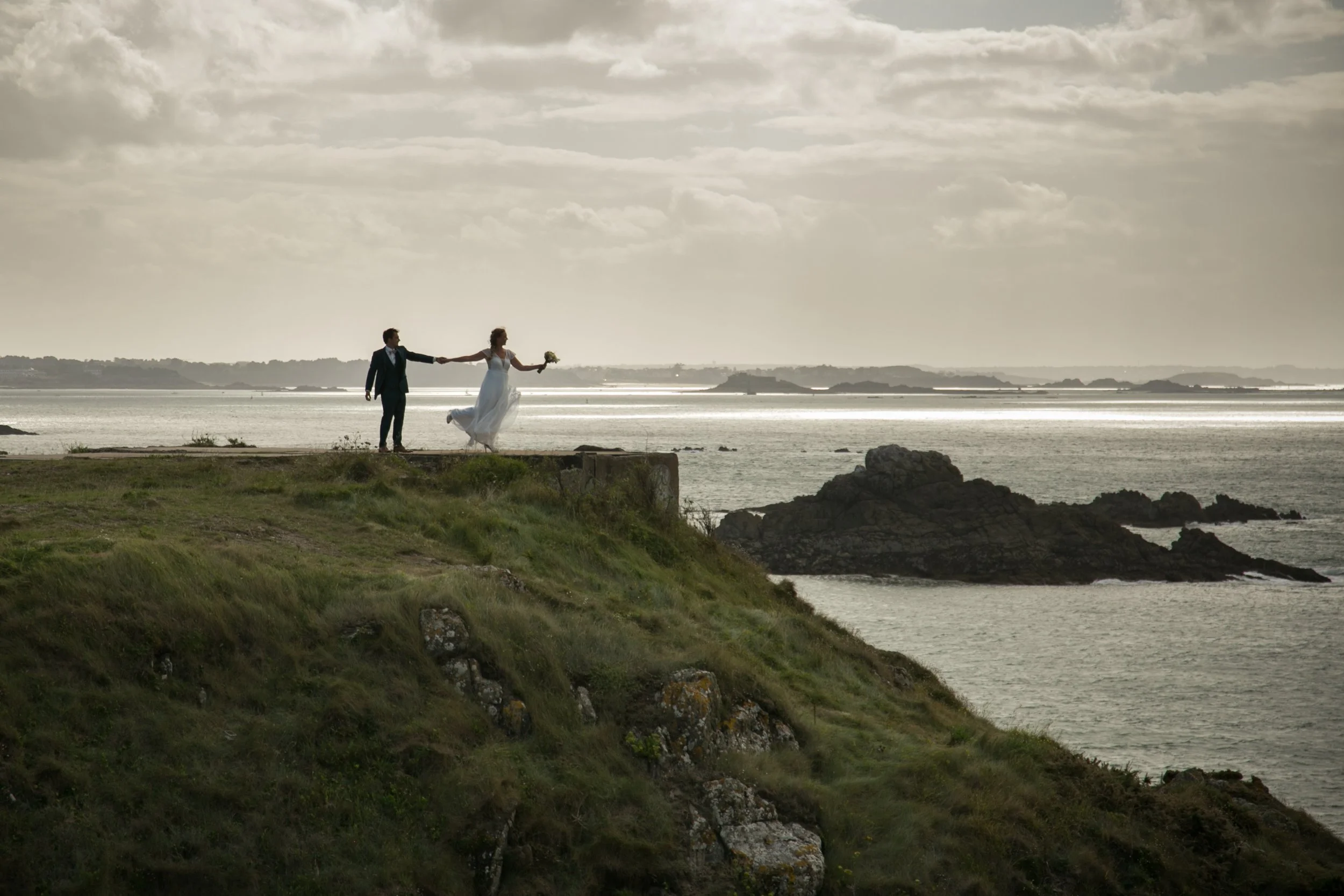 Un couple de mariés se tient sur une falaise au bord de l'eau, la femme tenant un bouquet de fleurs et le marié en costume noir, lors d'une scène de mariage en extérieur. saint-malo, Bretagne . casa19