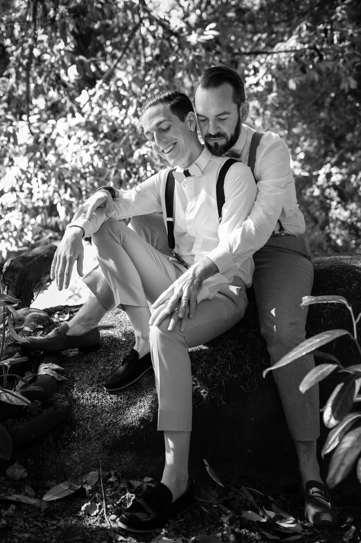 Deux hommes habillés en chemises blanches et pantalons, assis sur un arbre dans la nature, souriant et montrant une proximité affectueuse. L'un d'eux porte des bretelles.