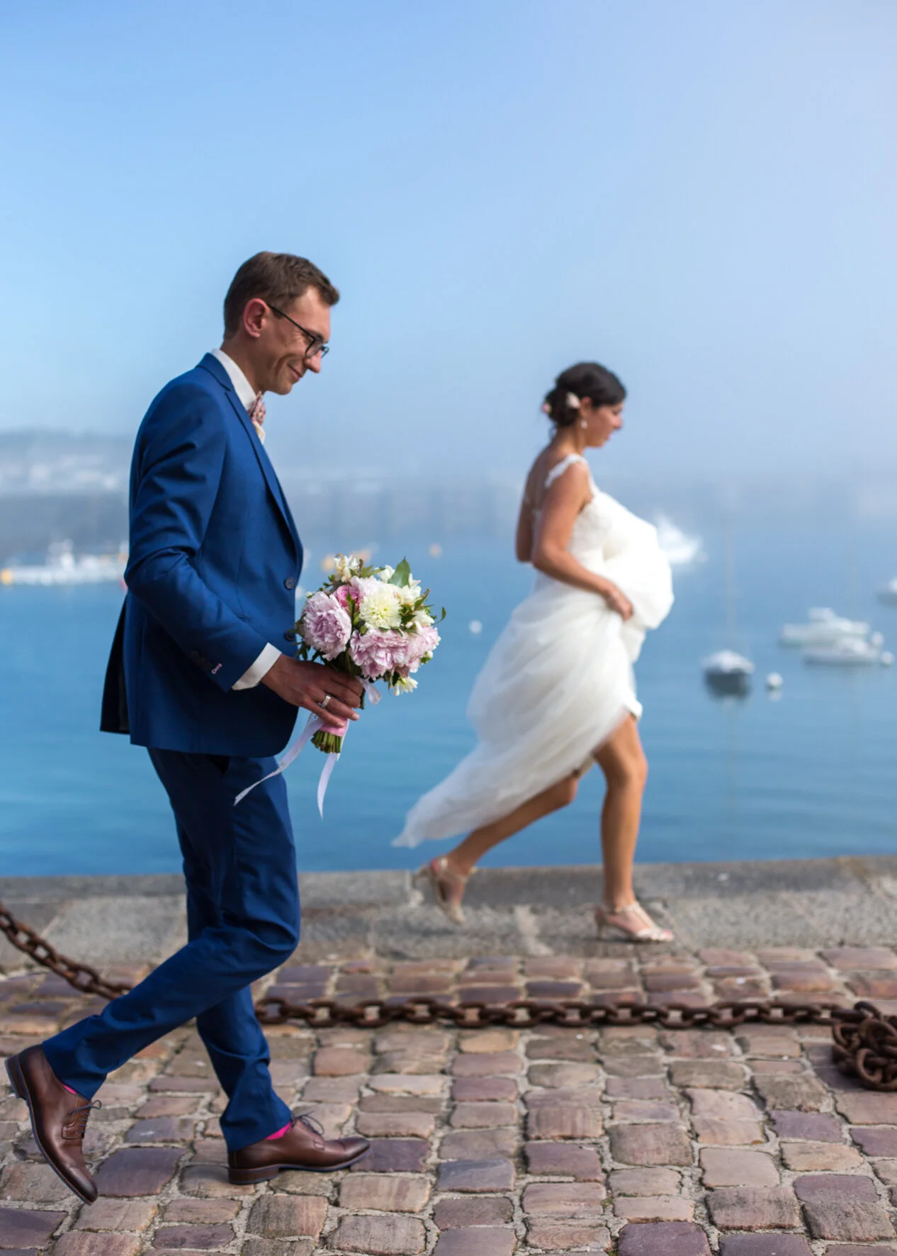 A groom in a blue suit holding a bouquet of pink and white flowers, walking along a cobblestone path near the water, with a pregnant bride in a white dress walking behind him. The background shows a body of water with boats and a clear sky.