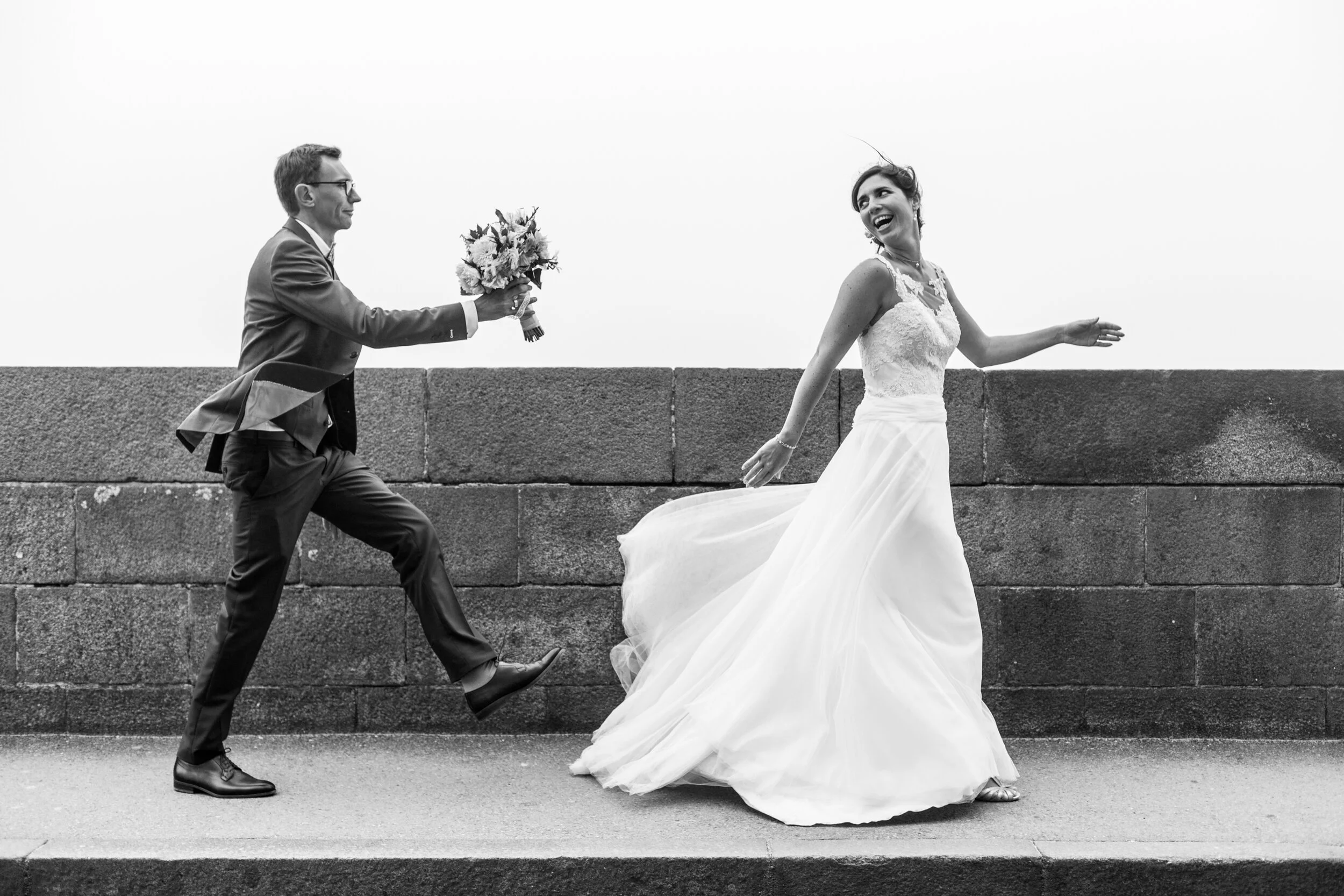 Black and white photo of a joyful bride in a wedding dress running away from a groom in a suit holding a bouquet of flowers, on a paved path with a stone wall in the background.