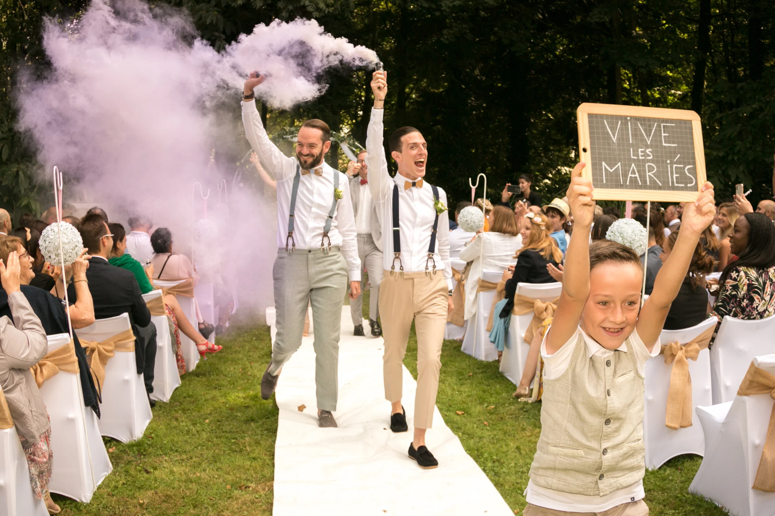 A joyful wedding celebration outdoors with two gay grooms walking down the aisle, one holding a smoke flare, surrounded by seated guests and a boy holding a sign that says 'VIVE LES MARIÉS.'