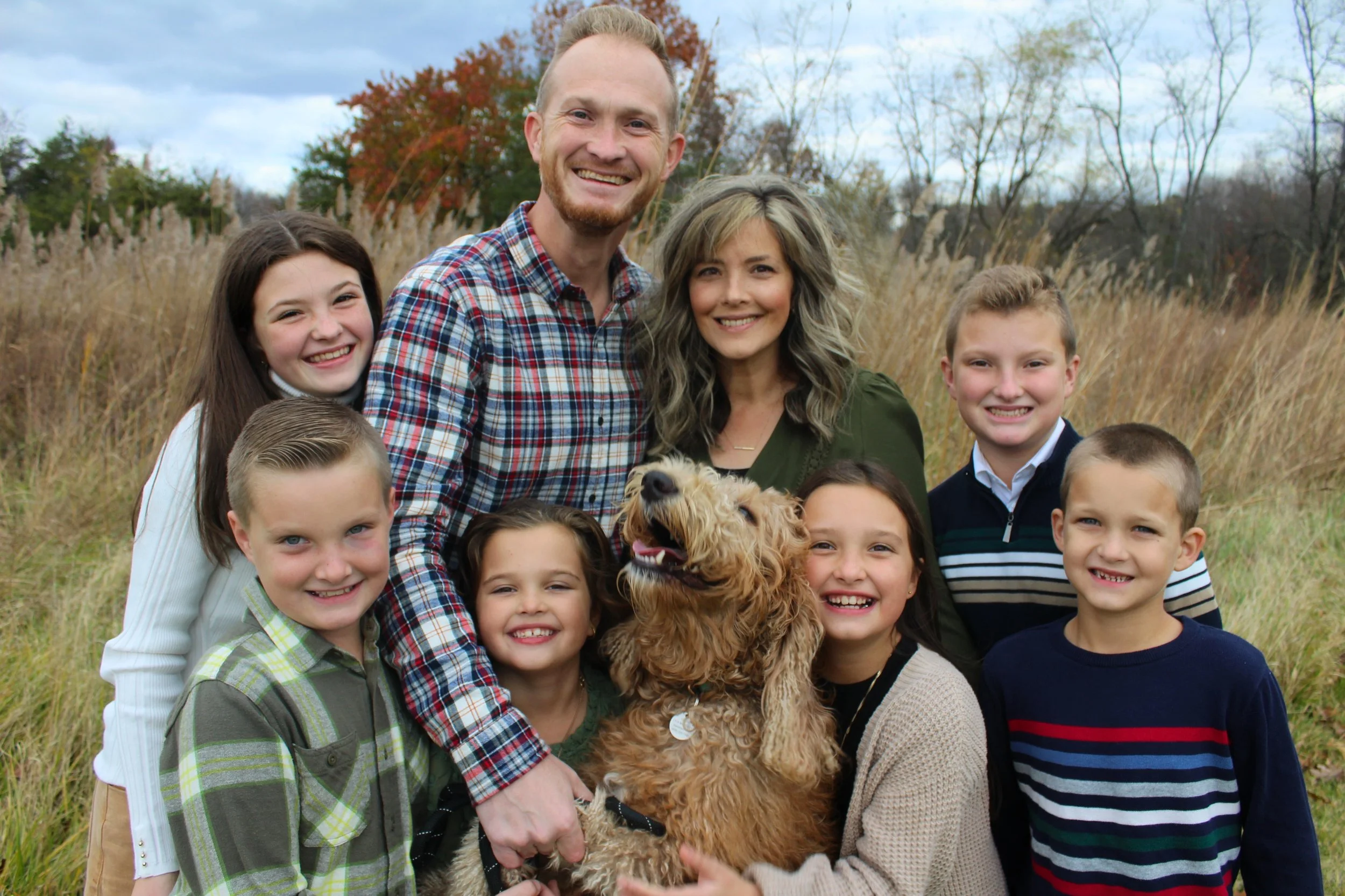 A group of children and two adults smiling outdoors in a field during fall, with trees in the background. One person holds a fluffy dog.