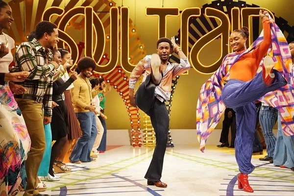 Two dancers in bright clothes kick into the air with other dancers lined up on the side in front of the words "Soul Train"