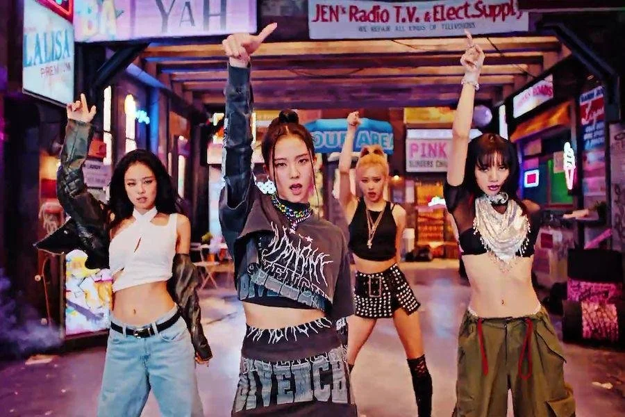 Four women in crop tops look at the camera and point upwards with their right hands