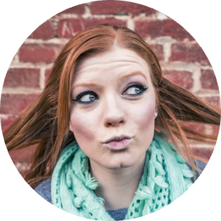 Woman with long red hair and blue scarf pulls her hair out to either side while looking to the right of the camera, eyebrows raised, making a kissing face