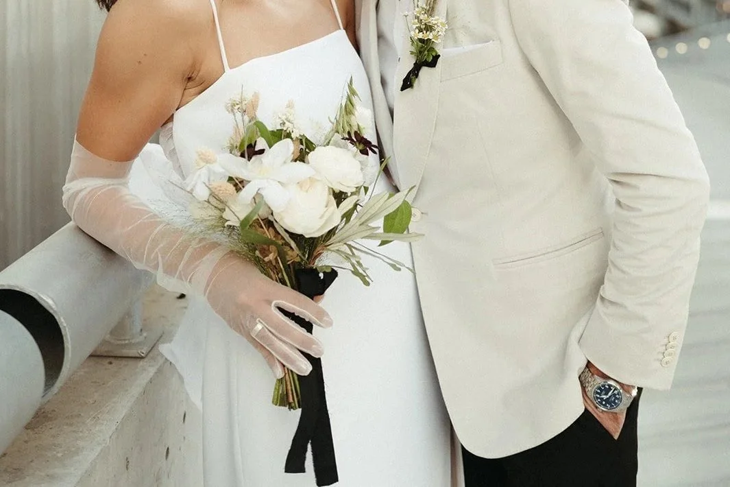 A bride and groom kissing outdoors, with the bride holding a bouquet of white flowers and the groom wearing a white suit jacket with a boutonniere, in an urban setting with modern buildings.