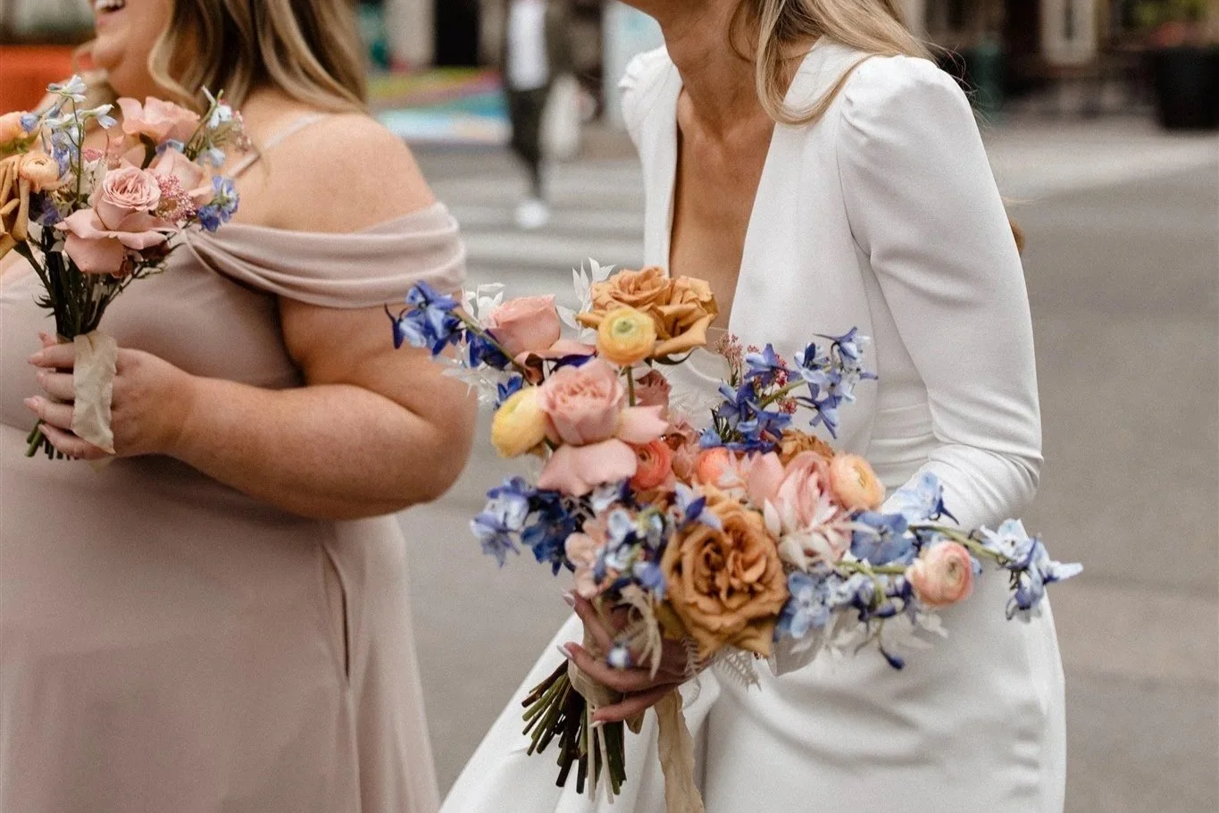 Two women in wedding dresses holding bouquets, laughing on city street. Floral arrangement by Foxglove Studio, Calgary.