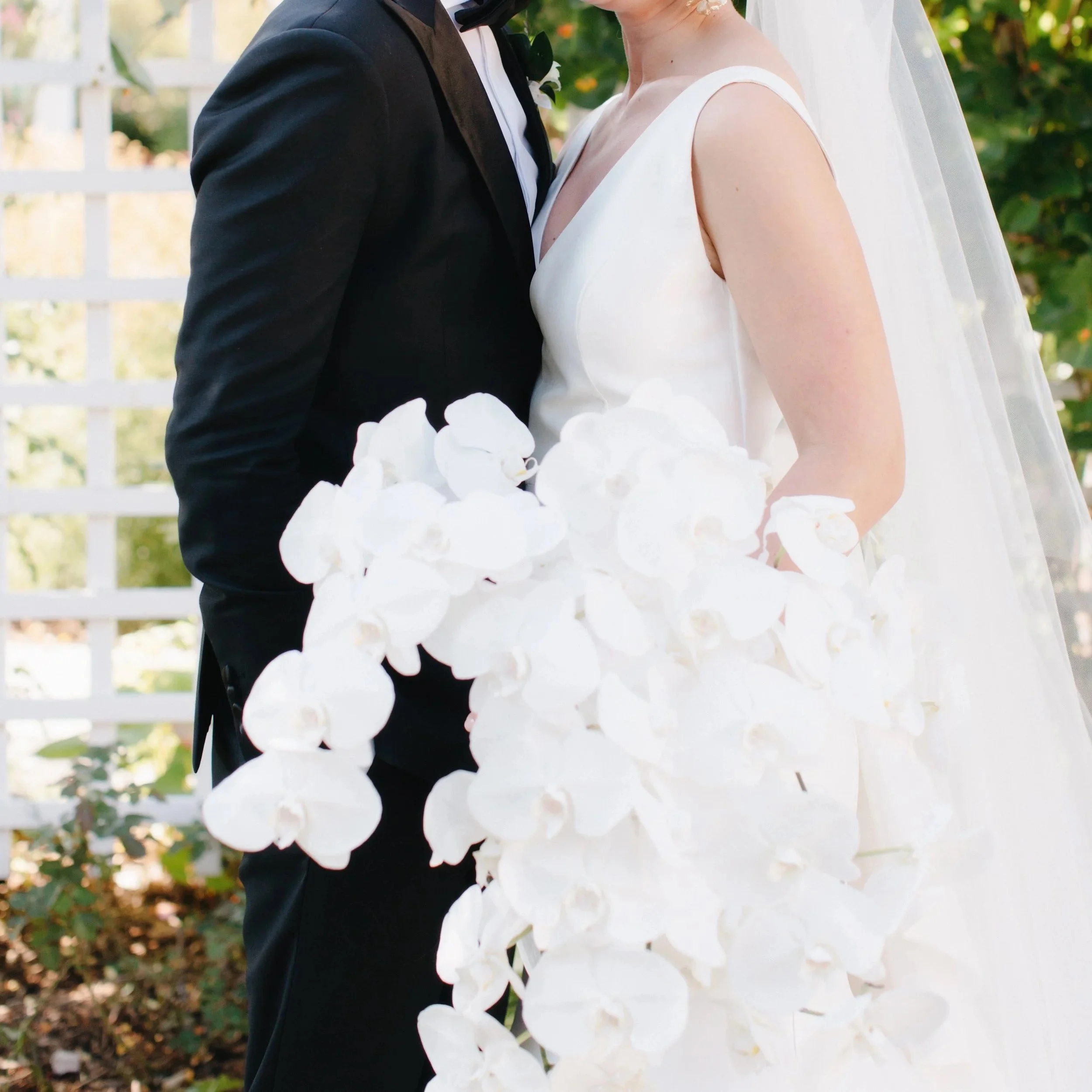 Bride and groom sharing a joyful moment outdoors. The bride is smiling, dressed in a white wedding gown with a veil and floral earrings, holding a large white flower bouquet. The groom is in a black tuxedo, whispering to the bride. Green foliage and 