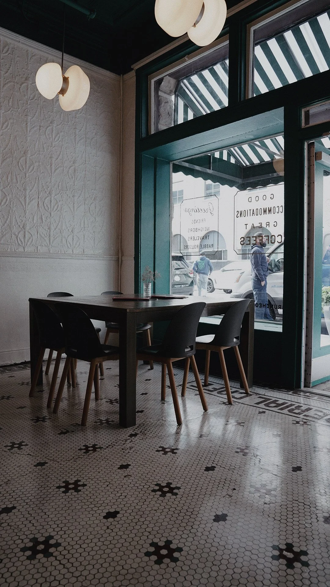 Interior of a cafe with a small table, four black chairs, patterned tile floors, large window with green trim, and white textured wall with hanging lights.